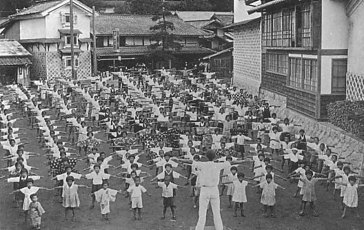 A 1930s Japanese schoolyard full of children doing radio calisthenics with an instructor leading